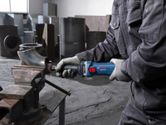 A person wearing safety equipment uses a straight grinder to grind a metal pipe in a workshop.