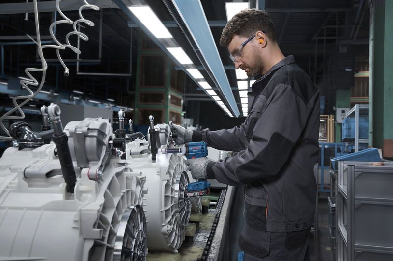 A person wearing safety equipment assembles machinery parts using a cordless power tool.