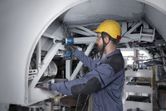 A person wearing safety equipment uses an impact wrench on a metal frame.