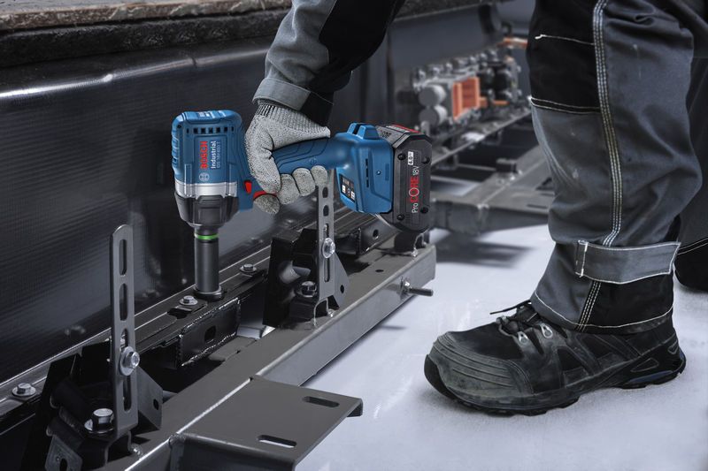 A person wearing safety equipment uses an impact wrench to fasten a bolt on a metal frame.