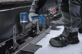 A person wearing safety equipment uses an impact wrench to fasten a bolt on a metal frame.