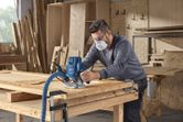 A person wearing safety equipment uses a router to shape wood in a workshop.