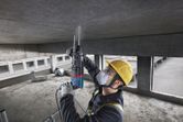 A person wearing safety equipment uses a rotary hammer to drill into a concrete ceiling.