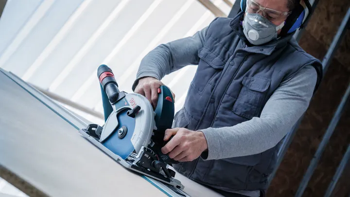 A person wearing safety equipment operates a circular saw to cut a sheet of wood.