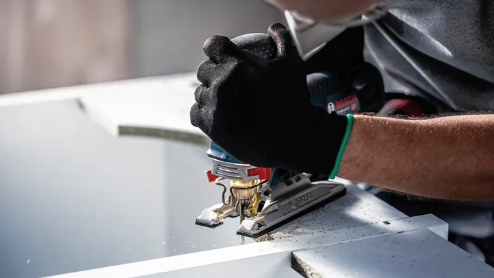 Person wearing safety equipment uses a jigsaw to cut a sheet of material.