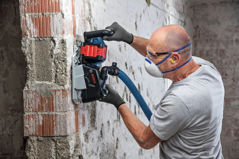 A person wearing safety equipment uses a wall chaser to cut grooves in a brick wall.