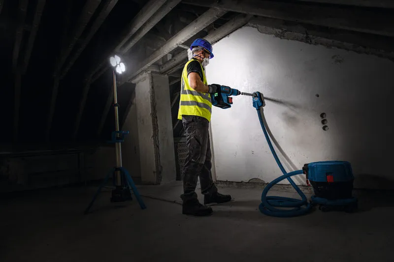 A person wearing safety equipment drills into a wall under a cordless jobsite light.