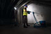 A person wearing safety equipment drills into a wall under a cordless jobsite light.