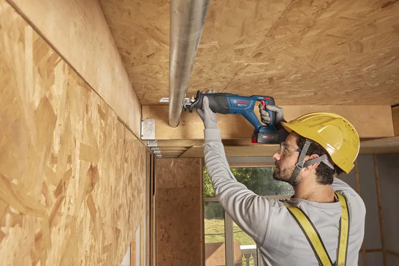 A person wearing safety equipment uses a cordless reciprocating saw to cut a ceiling pipe.
