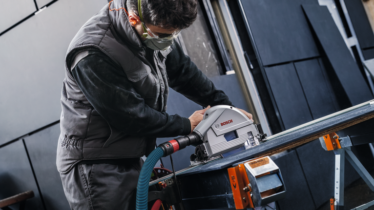 Person wearing safety equipment operates a circular saw to cut a sheet of material.