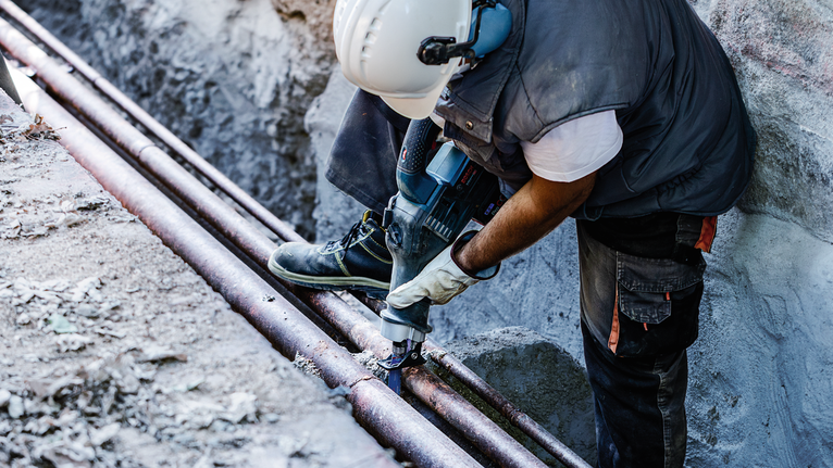 Person wearing safety equipment uses a power tool to cut metal piping outdoors.