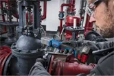 A person wearing safety equipment uses a cordless impact wrench on industrial piping.