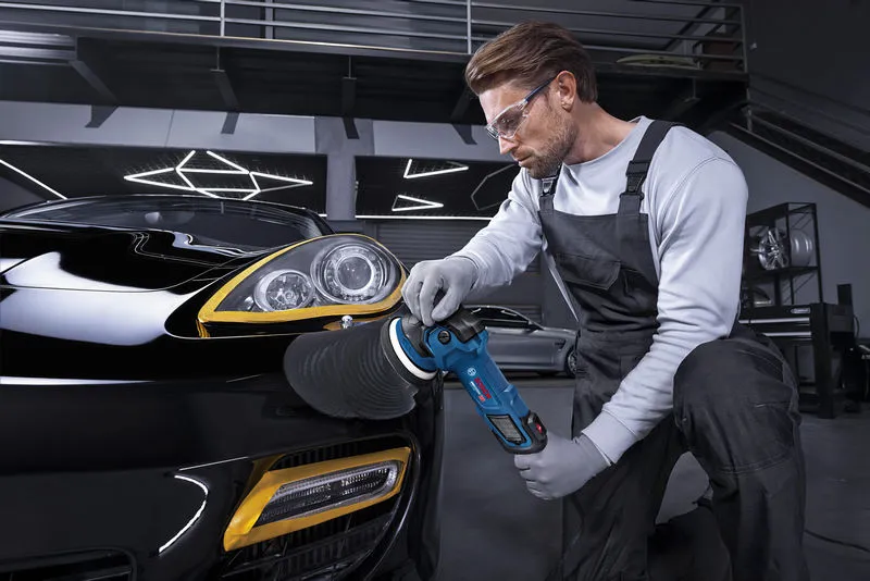 A person wearing safety equipment polishes a black car’s headlight with a polisher tool.