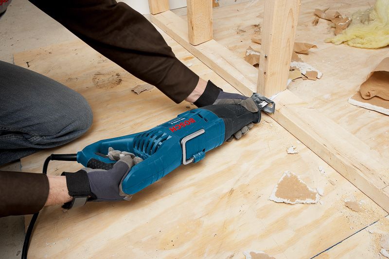 A person wearing safety equipment uses a reciprocating saw to cut wood framing.