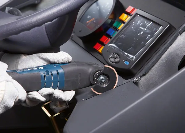 A worker wearing safety equipment uses a rotary tool to cut a dashboard panel.