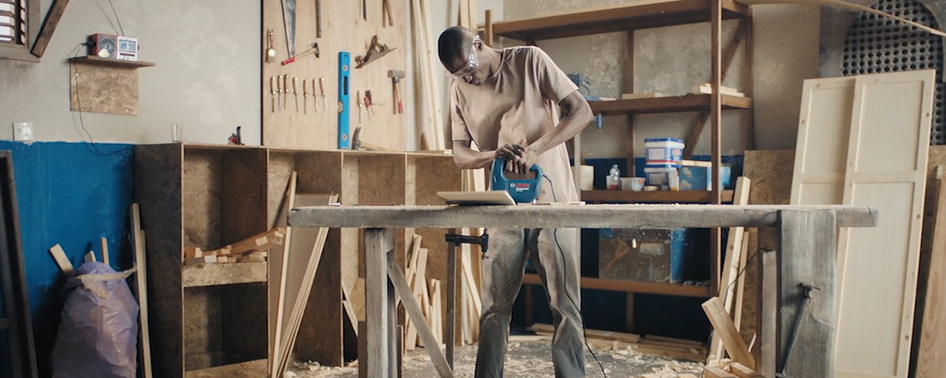 A man uses a blue Bosch jigsaw on a wooden workbench in a workshop filled with tools.